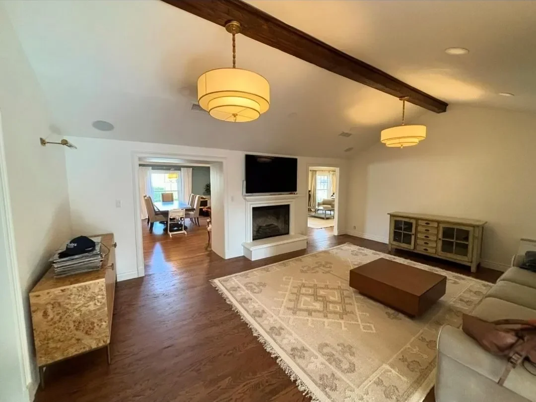 Living room with hardwood floors, a fireplace, a mounted TV above it, and a patterned area rug. There is a cream-colored sofa, a brown coffee table, and a sideboard. Two ceiling lights hang from a wooden beam, and an additional room is visible throug