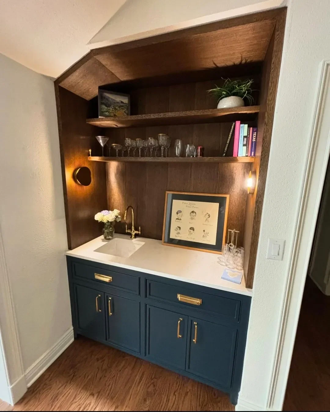 A home bar area with a blue lower cabinet, a white countertop with a small sink, a gold faucet, and shelves with decorative items and glassware, illuminated by wall-mounted lights.