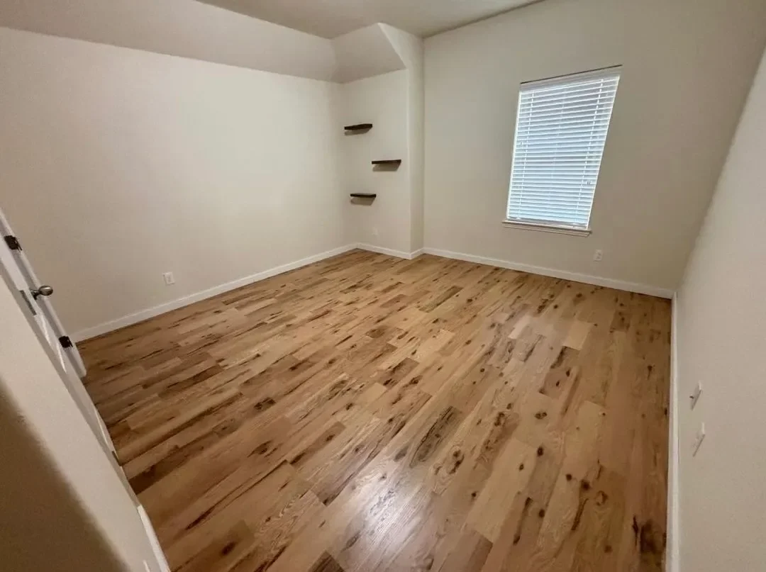 Empty room with hardwood floors, white walls, a window with blinds, and three small wall-mounted shelves in the corner.