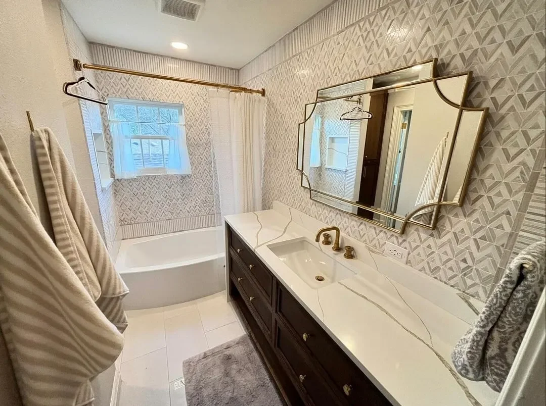 A bathroom with a bathtub, a marble vanity with a sink and gold fixtures, a decorative mirror, beige and patterned wallpaper, a window with a curtain, towel racks with towels, and a gray rug.