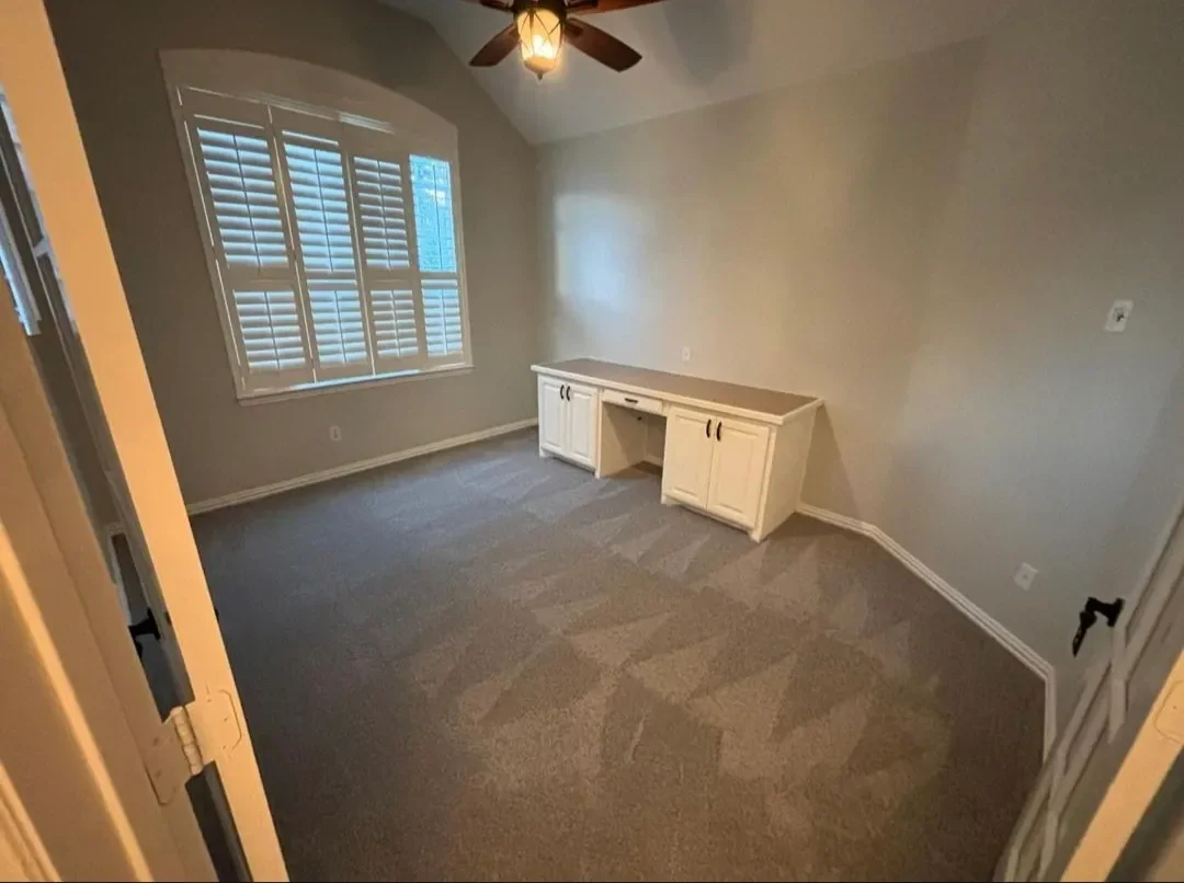 Empty room with beige carpet, light-colored walls, a ceiling fan, window with white shutters, and a white built-in desk.