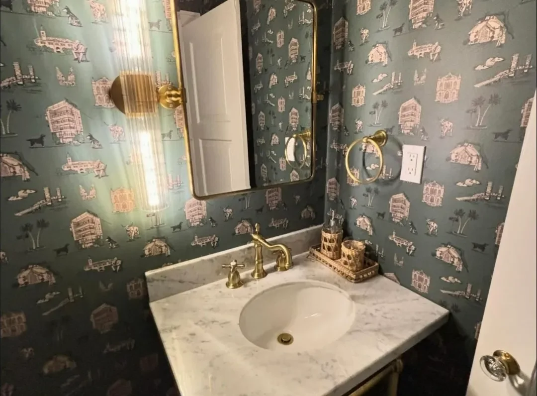 Bathroom sink with a marble countertop, gold faucet, decorative containers, a mirror, gold towel ring, and wallpaper with images of houses, trees, animals, and other scenery.