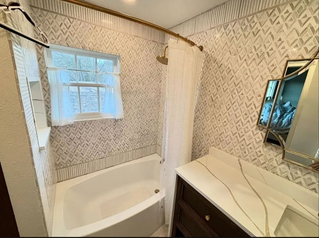 Bathroom with a bathtub, showerhead, window with curtains, mirror, sink with a marble countertop, and patterned wallpaper.