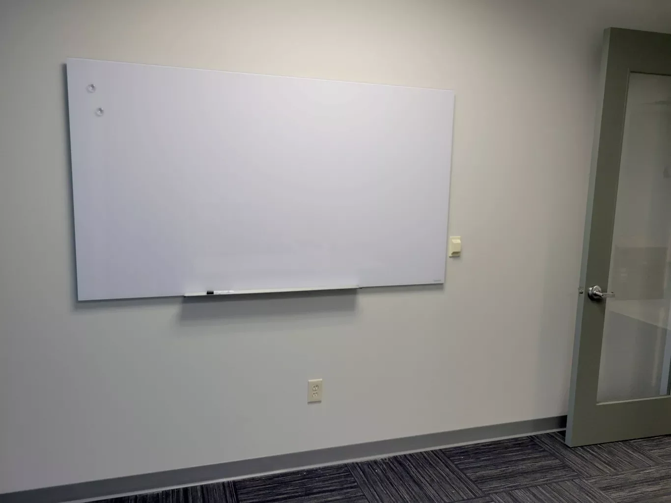 Empty whiteboard on a plain wall in an office or classroom, with a door to the right and electrical outlets below.