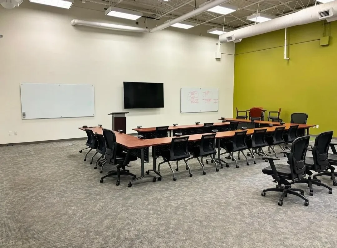 Empty conference room with a U-shaped table, black office chairs, whiteboards, a large monitor, and a green accent wall.