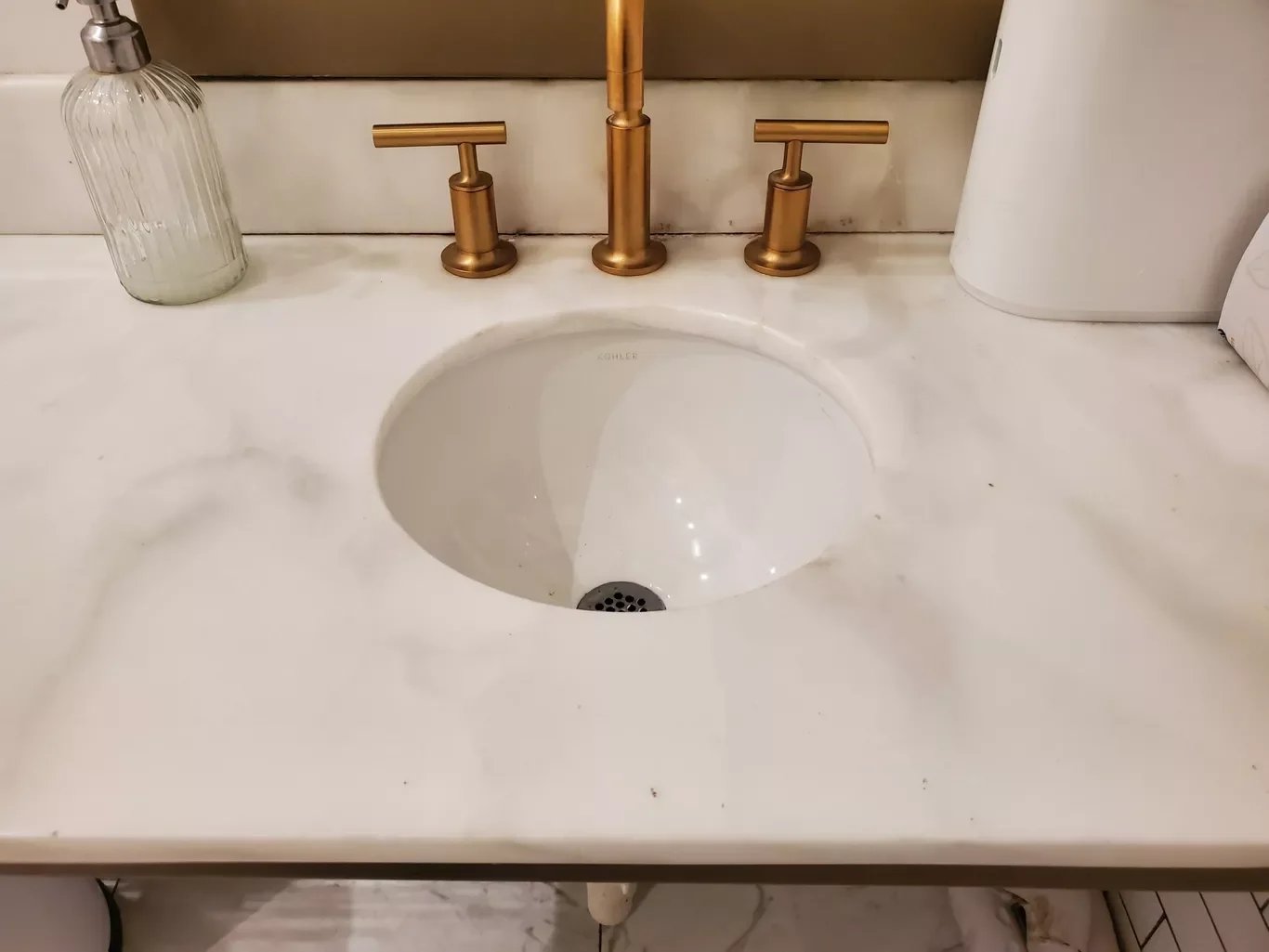 Bathroom sink with gold-colored faucet and handles, a clear soap dispenser on the left, and a white tissue box on the right, installed on a white marble countertop.