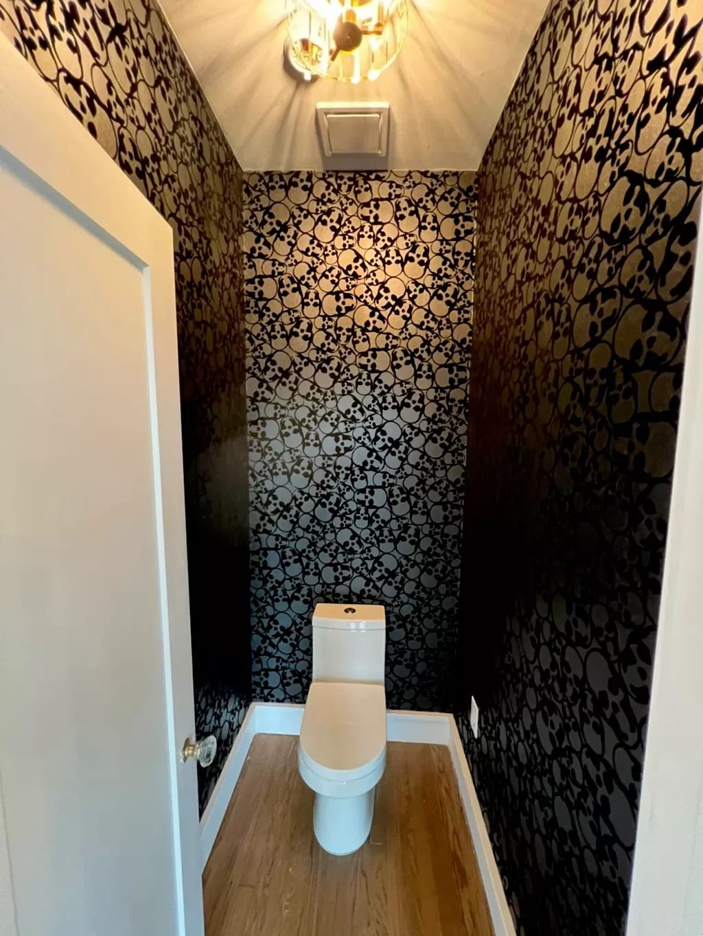 Small bathroom with black wallpaper featuring skull and floral patterns, a white toilet, wood flooring, and a ceiling light fixture.