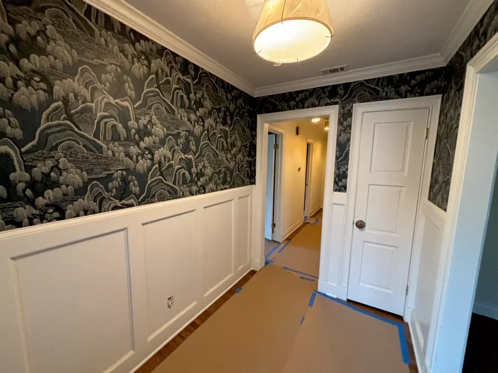 Hallway with black and gray patterned wallpaper, white wainscoting, white doors, a light fixture on the ceiling, and protective covering on the floor for painting or renovation.