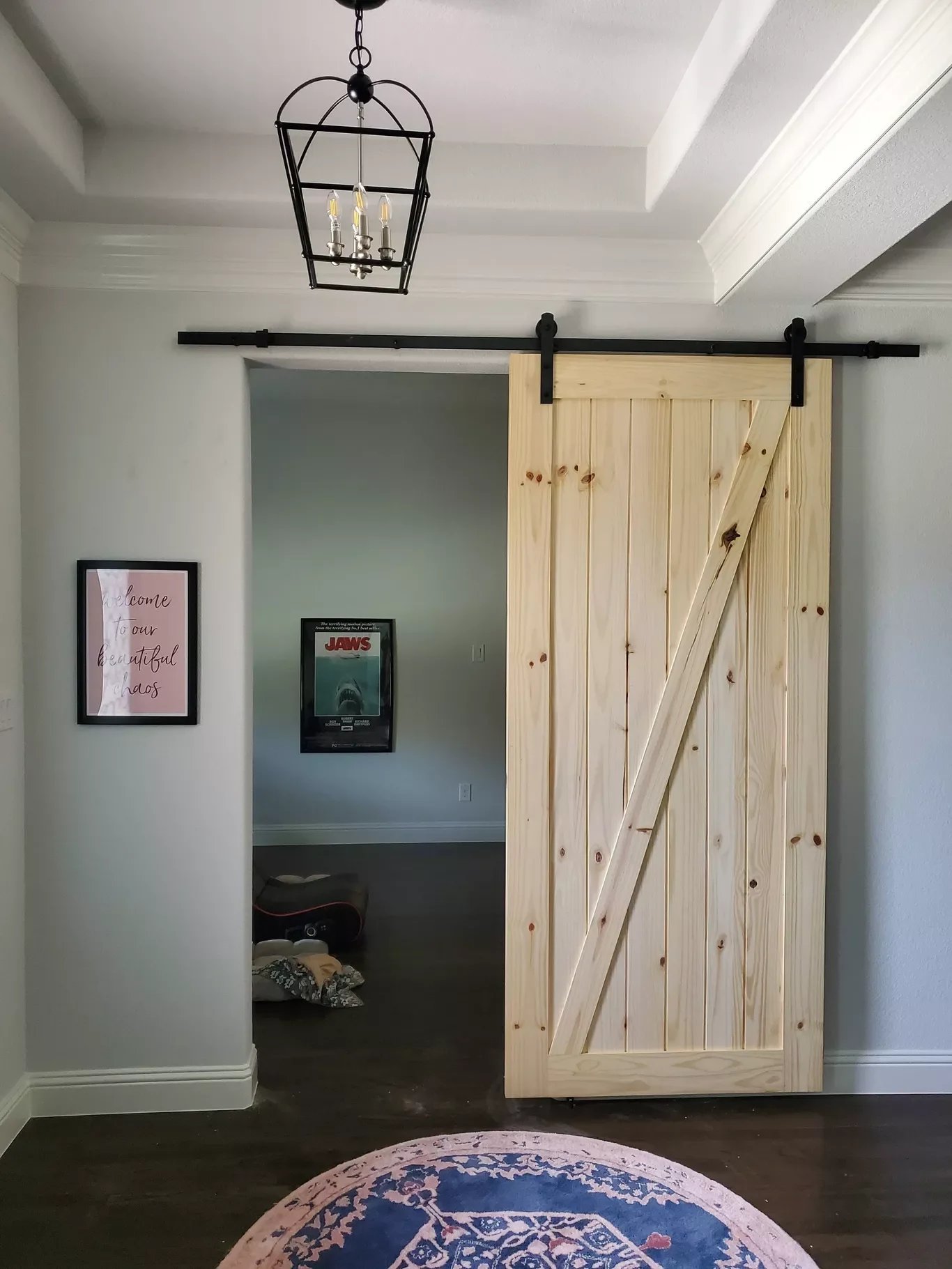 A partially open sliding barn door made of light wood on a black metal track, revealing a room with framed posters on the wall, a guitar case on the floor, and a pink and blue rug on dark hardwood flooring.