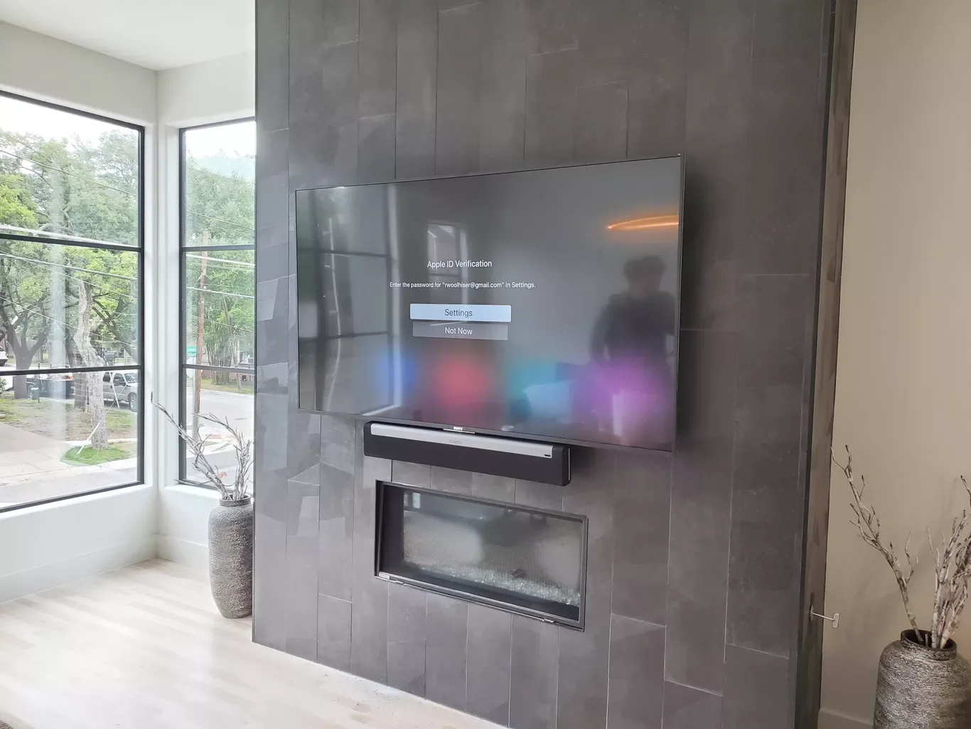 Smart TV on a gray tiled wall displaying Apple ID verification screen. A soundbar is mounted below the TV. Large windows with a view of trees and street outside, with a vase and decorative branches on the beige floor.