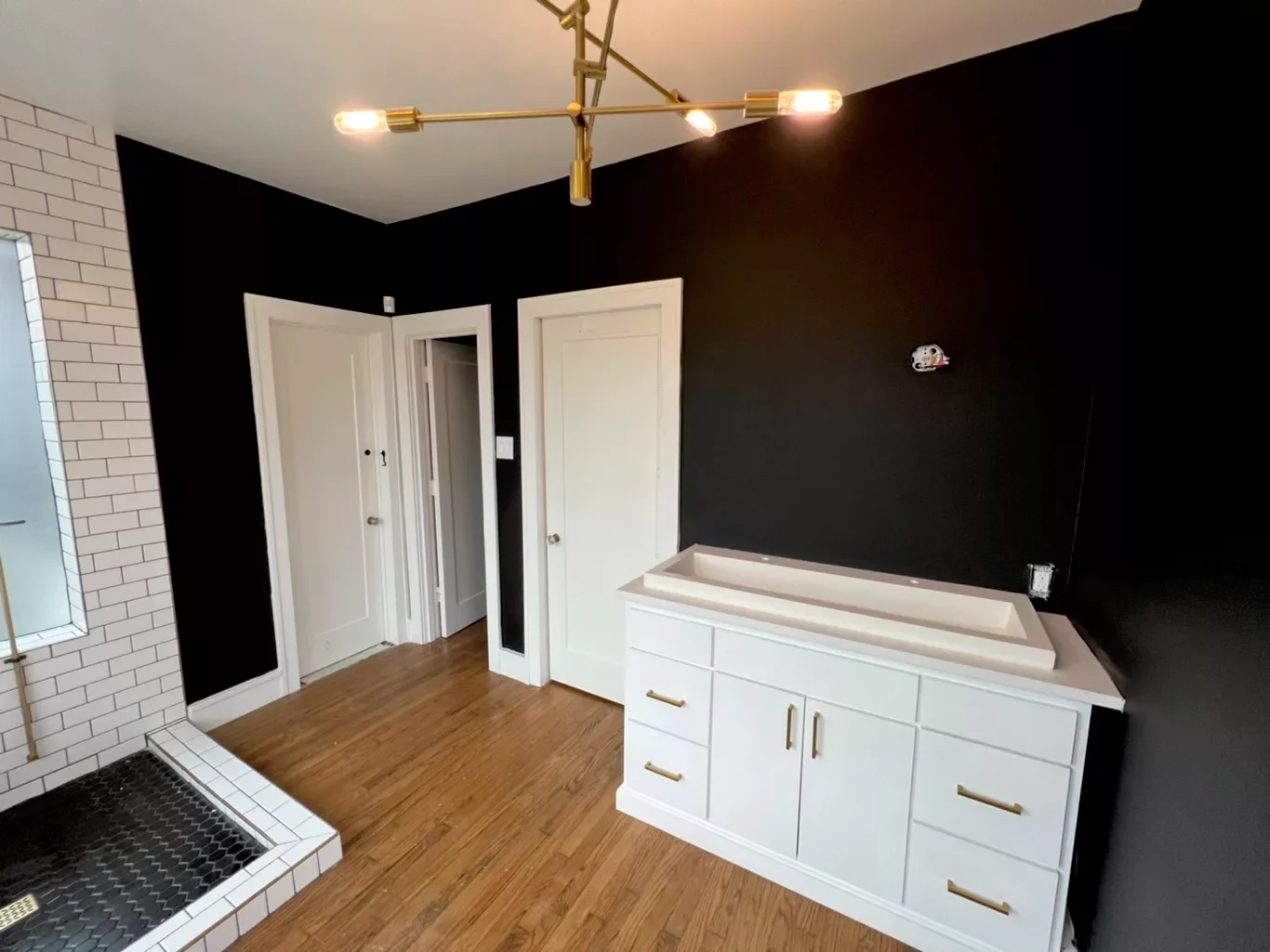 A modern bathroom with a wood floor, black and white walls, a white vanity with a sink, and a brass chandelier.