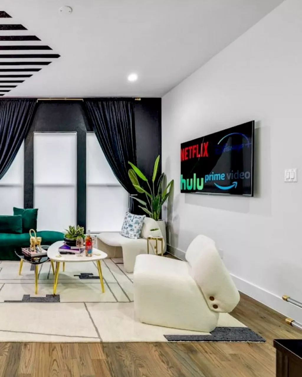 Modern living room with a wall-mounted TV displaying streaming service logos, a white sofa with patterned pillows, a potted plant, a small white coffee table with decorative items, a green velvet sofa, and black curtains over large windows.