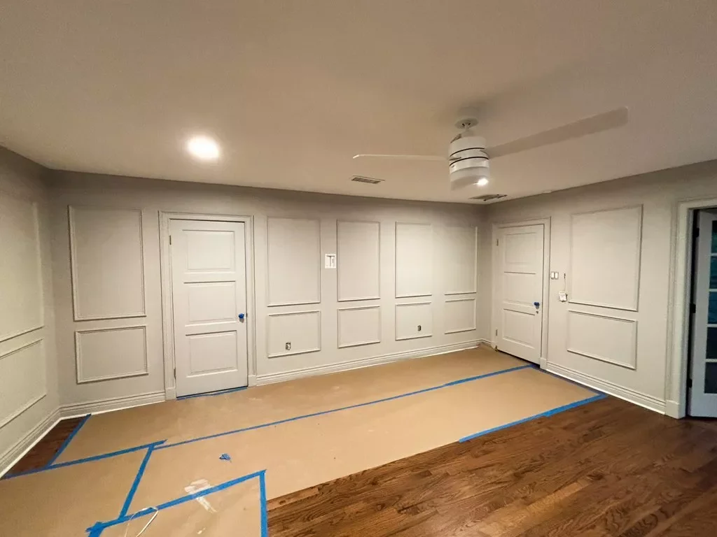 An empty room with white paneled walls, wooden floor, and doors, with masking tape on the floor indicating a pending renovation or assembly.