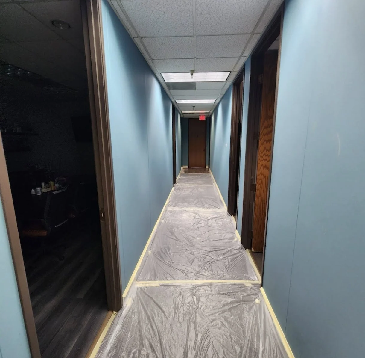 Empty hallway with blue walls, brown door at the end, and plastic covering on the floor, possibly for painting or renovation.