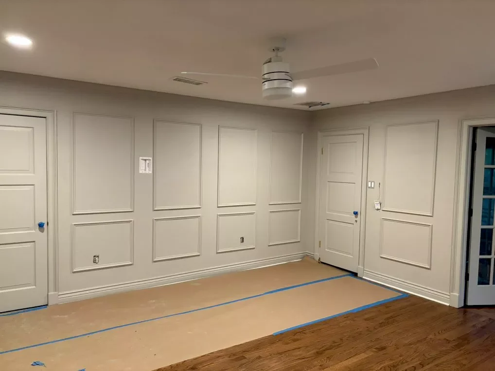 An empty room with beige walls, hardwood flooring, two closed white doors, and a ceiling fan with lights. The floor area is taped off with blue painter's tape, indicating ongoing work or renovation.