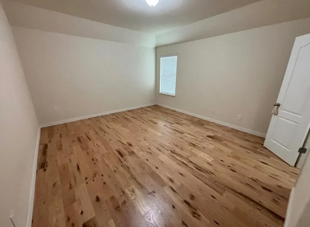 Empty room with hardwood floors, white walls, a window with blinds, and a closed white door.