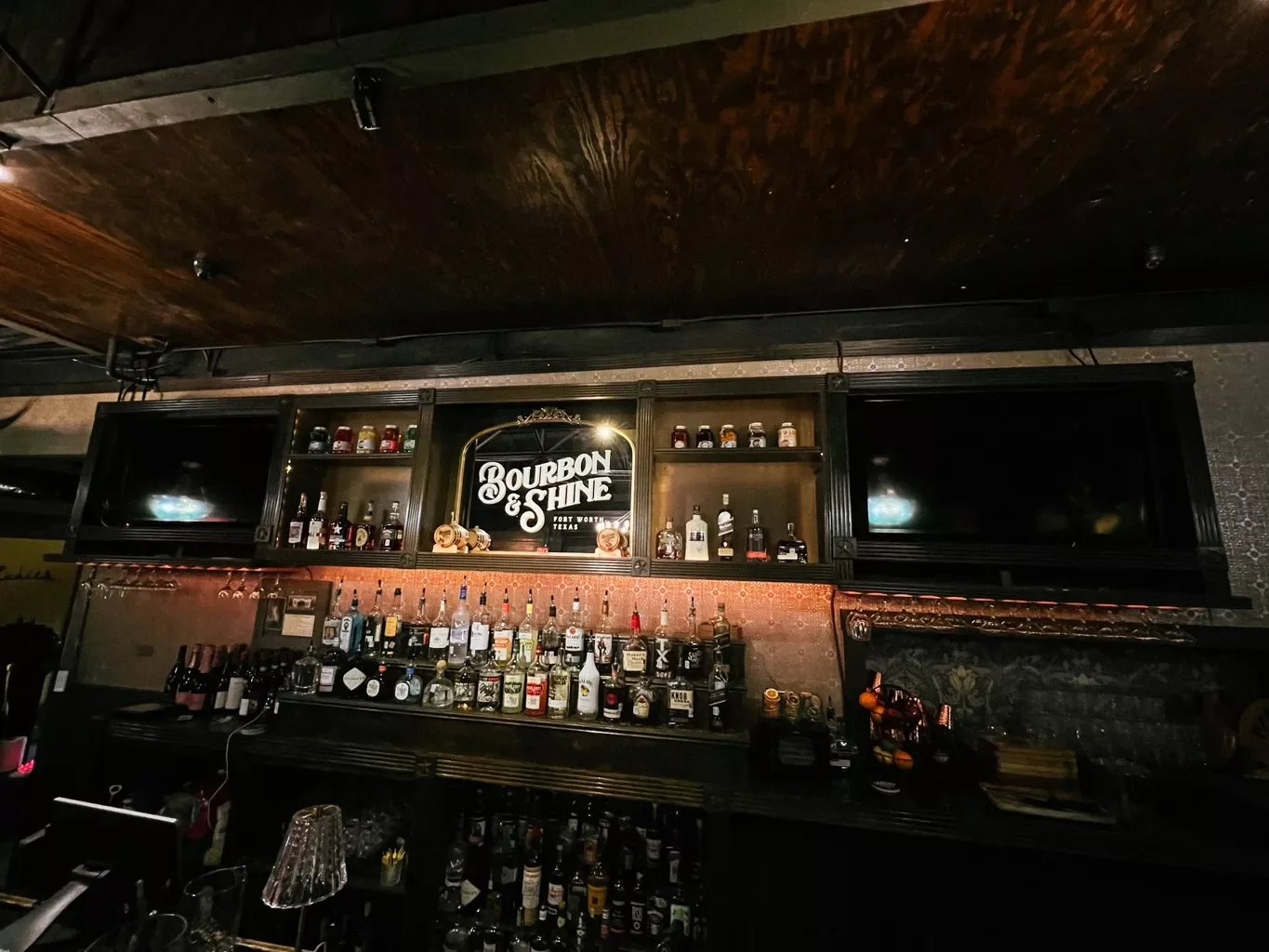 Bar with liquor bottles, shot glasses, and a illuminated sign that reads 'Bourbon & Shine' in a dimly lit setting.