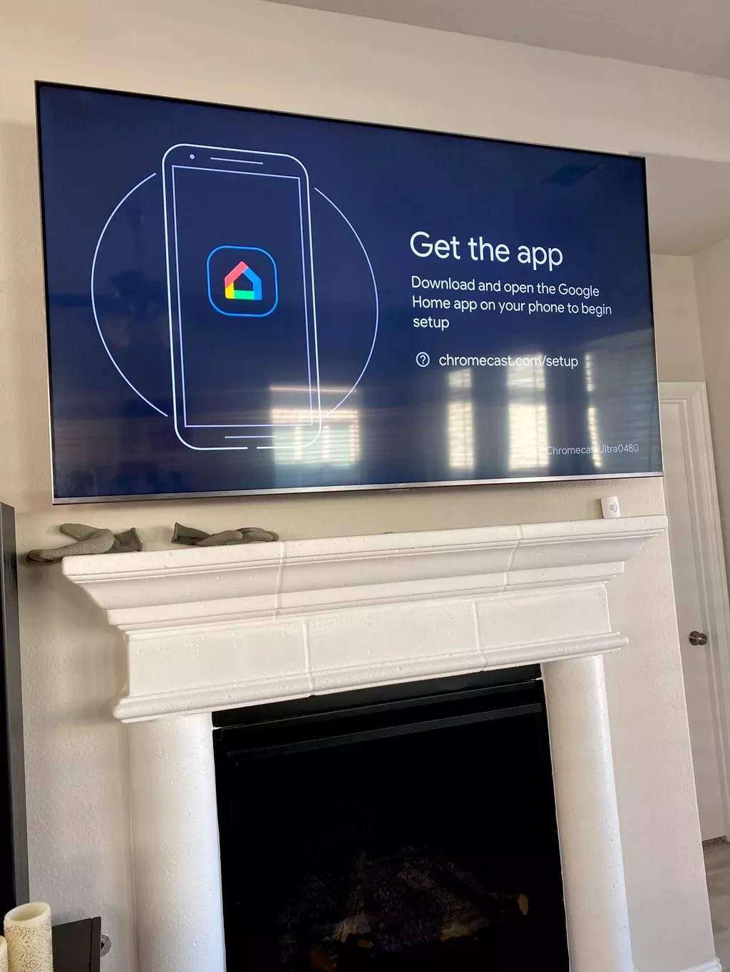 TV screen displaying setup instructions for Google Home app with a phone icon and colorful logo, mounted above a white fireplace mantel.