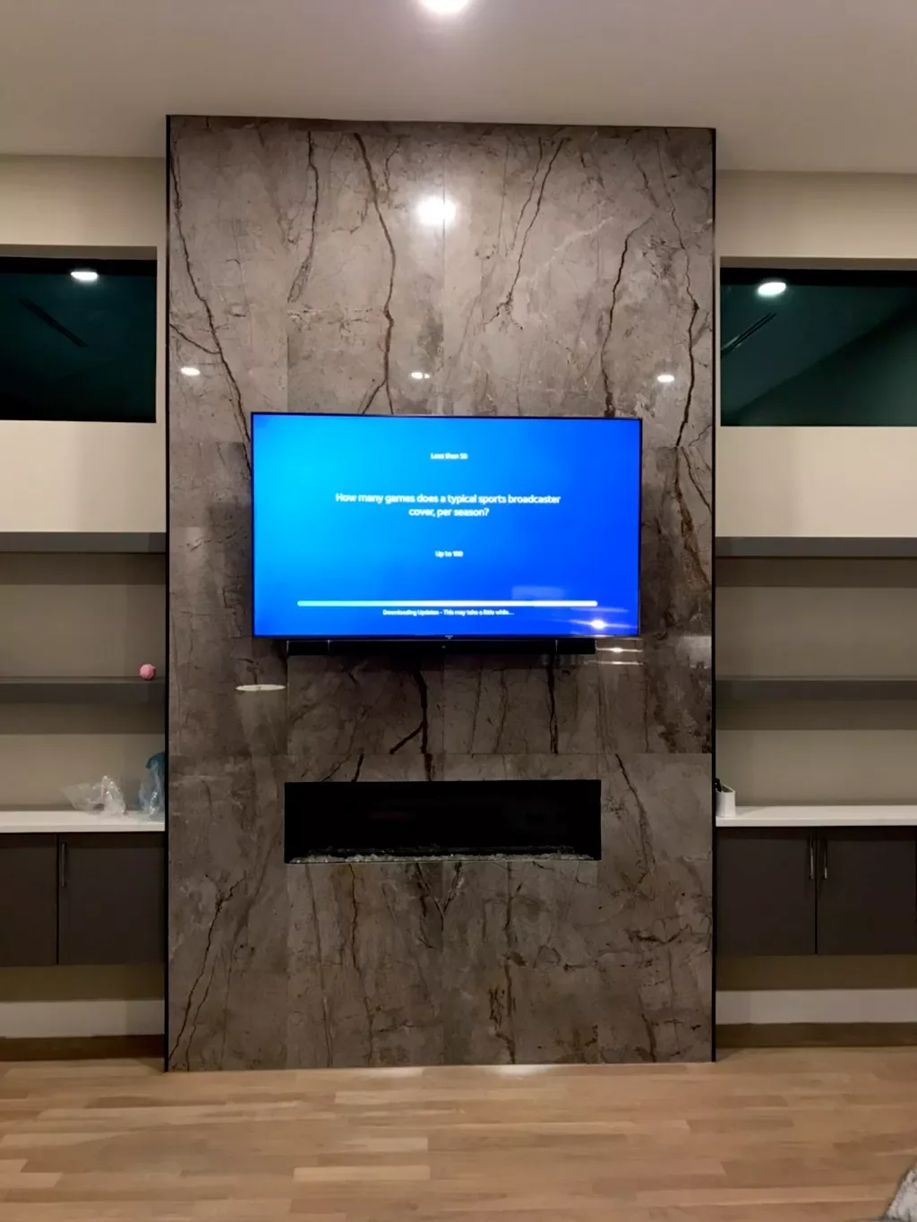 Television screen displaying a blue screen with a question about sports broadcasters, mounted on a marble-patterned wall with a built-in fireplace below, flanked by open shelves with various items in a modern room.