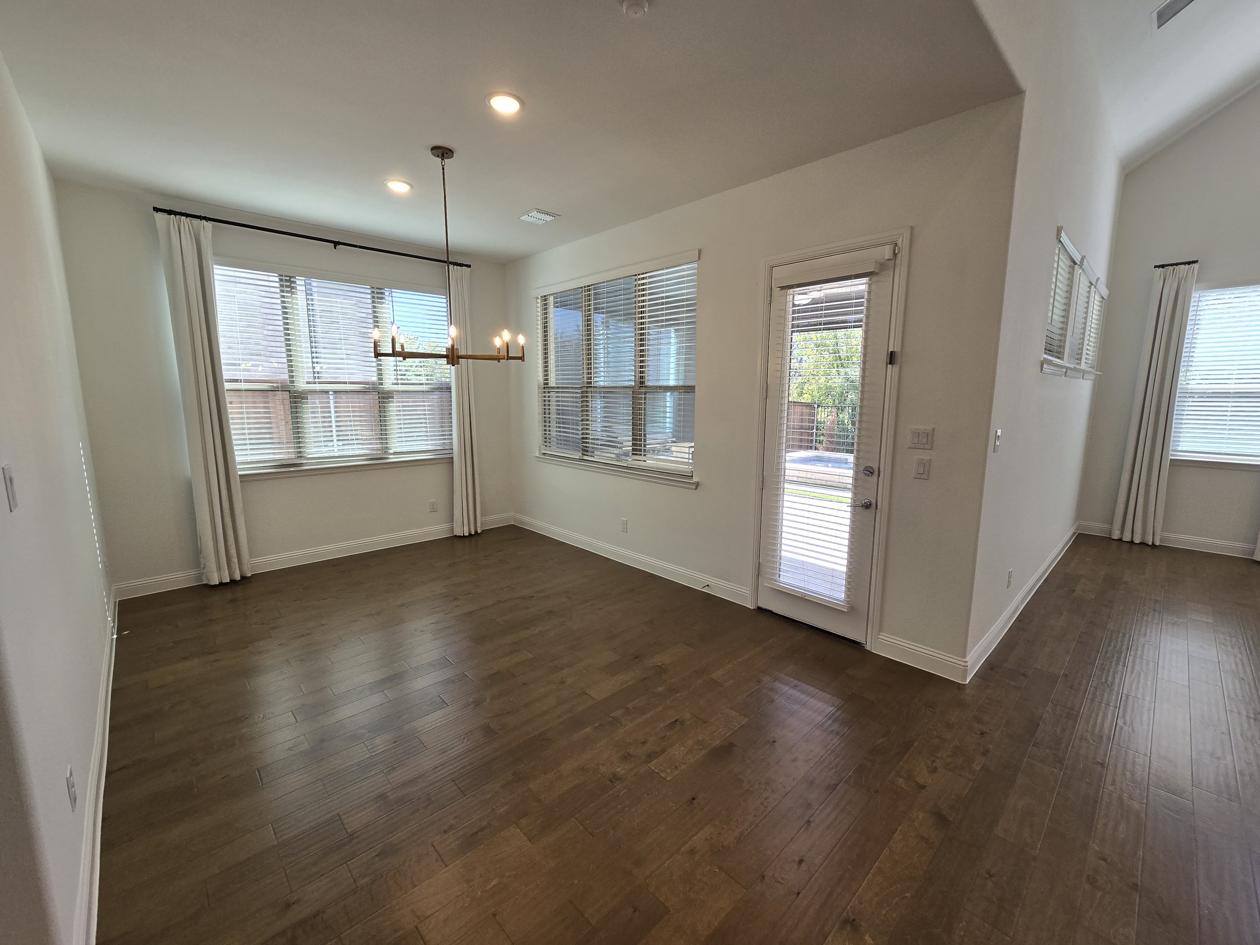 Empty living room with hardwood floors, white walls, large windows with blinds, and a chandelier hanging from the ceiling.