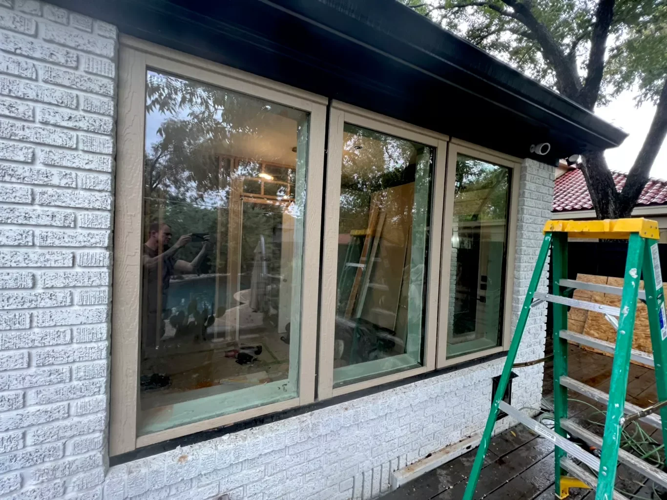 Construction site with a large sliding glass window, a green and yellow ladder, white brick wall, and a tree with a red-tiled roof in the background.