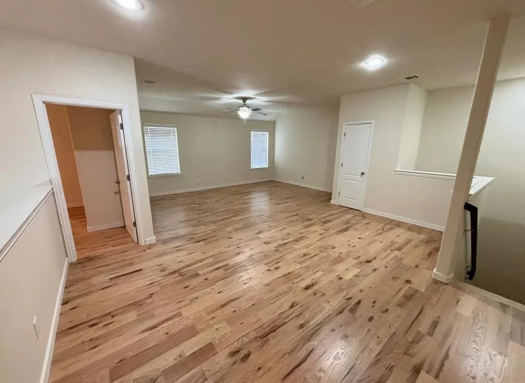 Empty living room with light-colored hardwood floors, white walls, and a ceiling fan with lights. Two windows with blinds are visible, and there is a small closet door and an open doorway leading to another room.