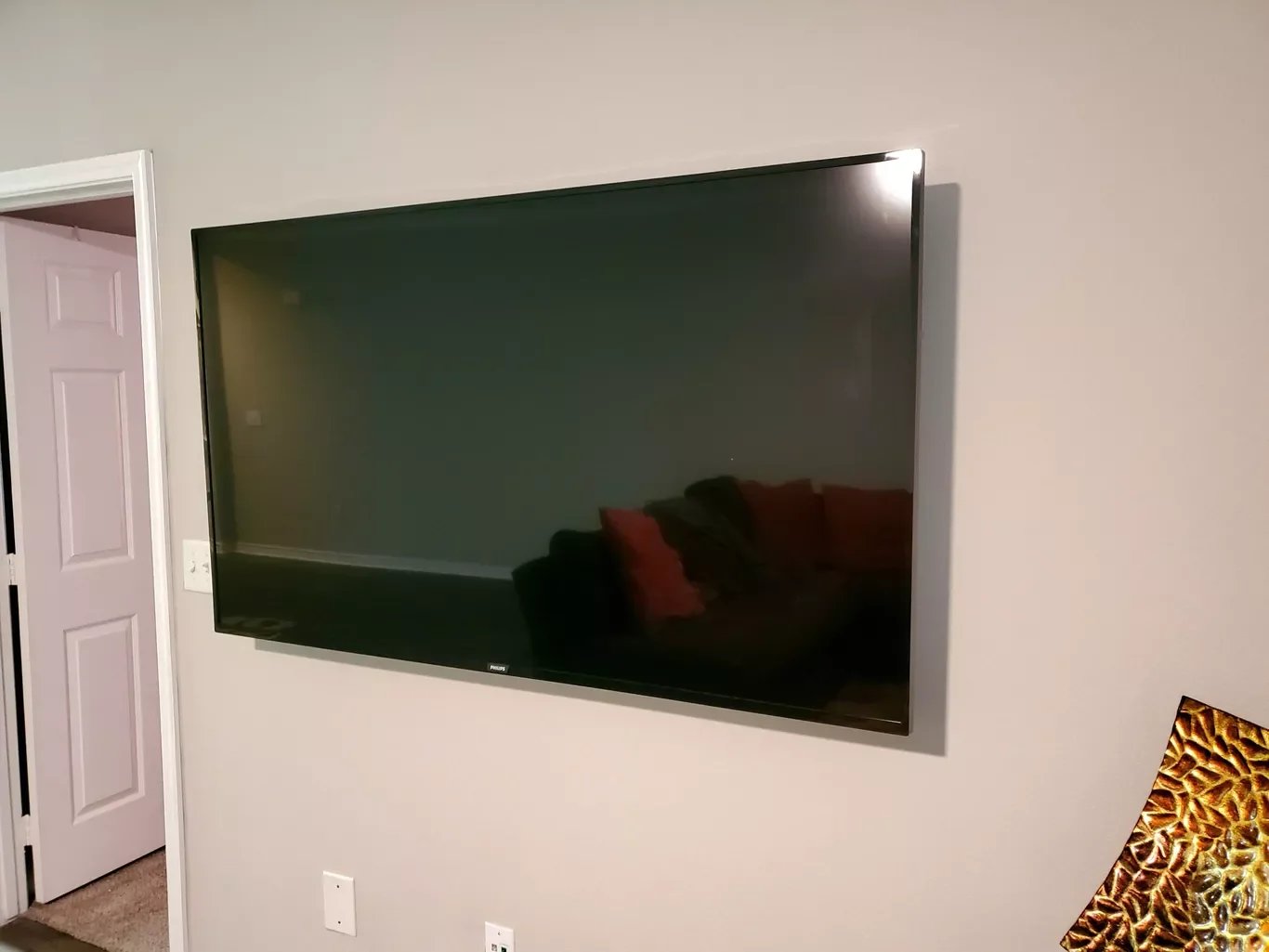 A large flat-screen television mounted on a beige wall in a living room, with a reflection of a couch with red and black pillows visible on the screen.