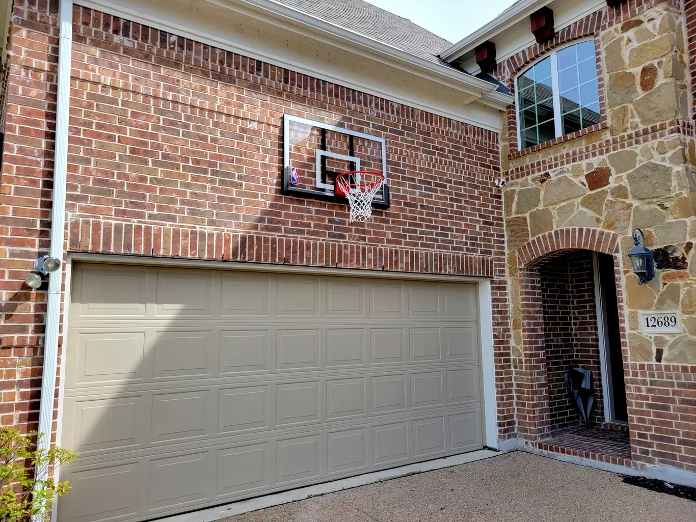 A basketball hoop mounted above a garage door on a brick house, with a lantern-style light fixture next to the entrance and a house number 12689.