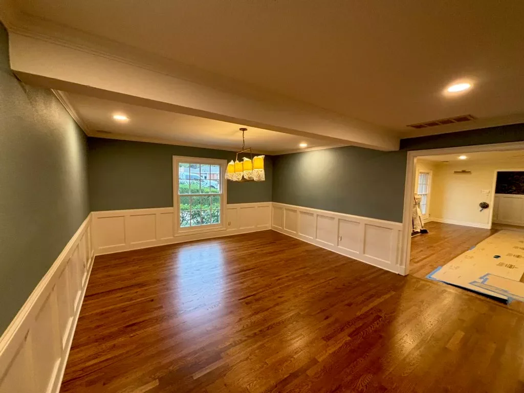 Empty dining room with hardwood floors, green walls, white wainscoting, window, and ceiling light fixture, connected to another room with light-colored walls.