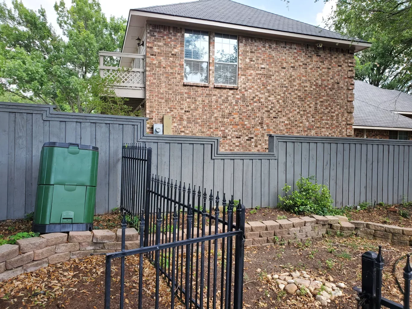 Backyard with a gray wooden fence, a small gate, a green trash bin, a brick garden bed, and a two-story brick house with a balcony and a window, surrounded by trees.