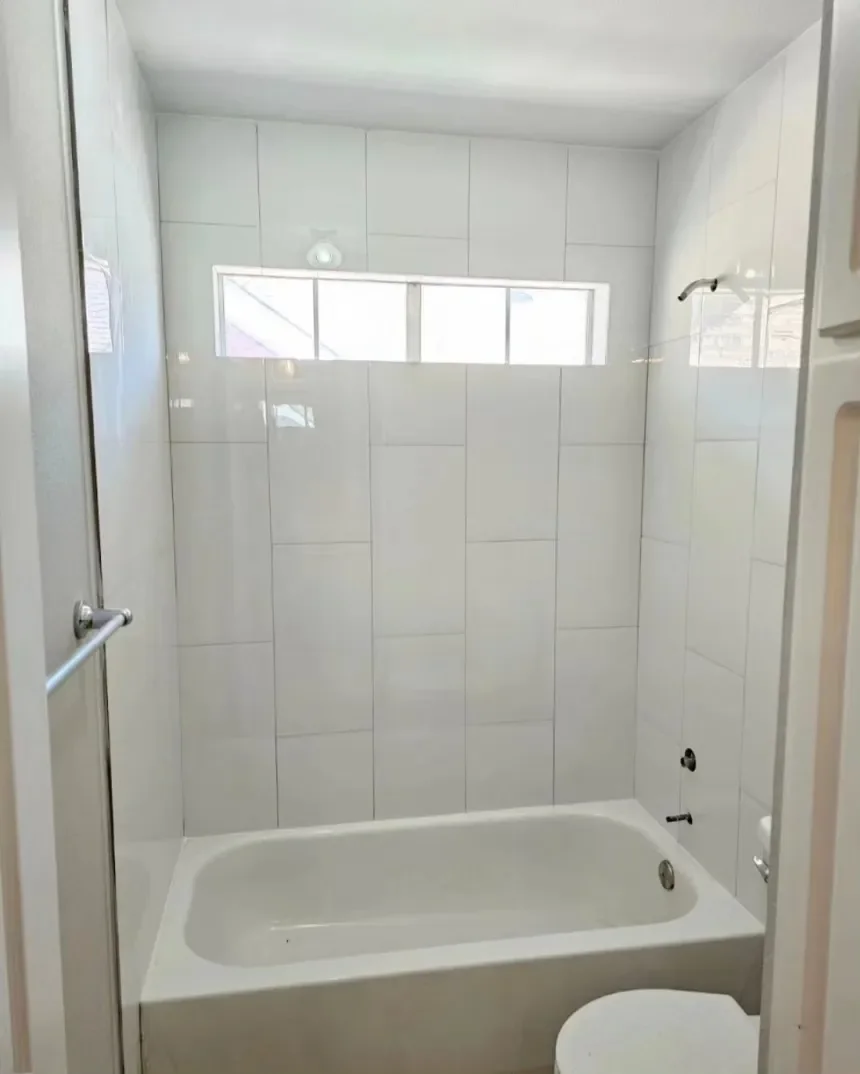 Empty bathroom with white tiled walls, a bathtub, a small horizontal window, and a toilet in the bottom right corner.