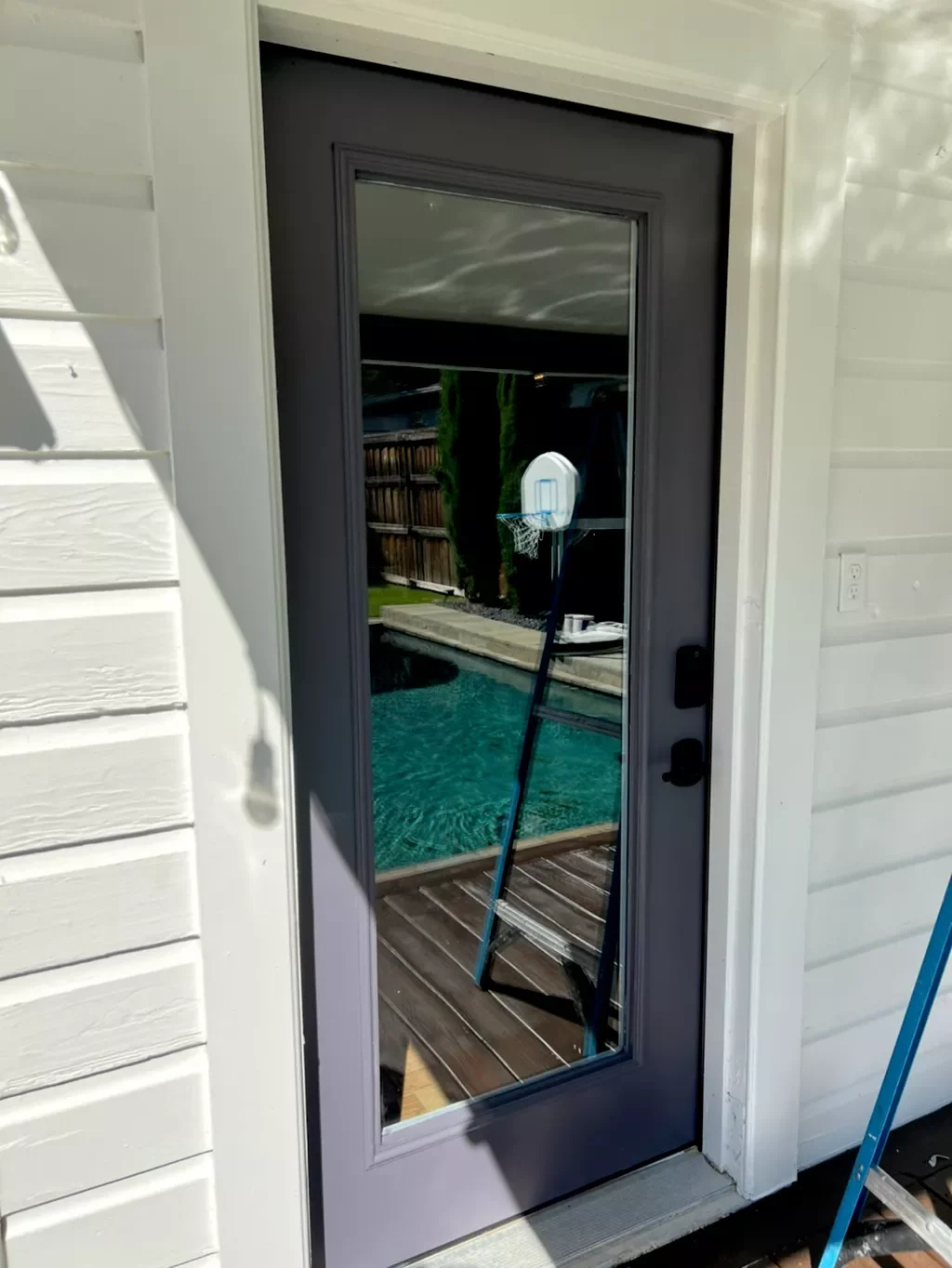 Reflective glass door showing pool, wooden deck, basketball hoop, and backyard trees.