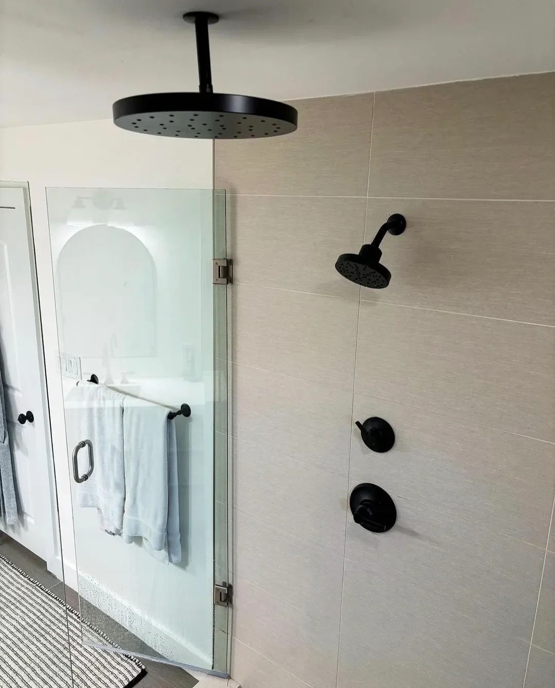 Modern shower area with two black shower heads on a beige tiled wall, a glass shower door, and a towel hanging on a bar inside the bathroom.