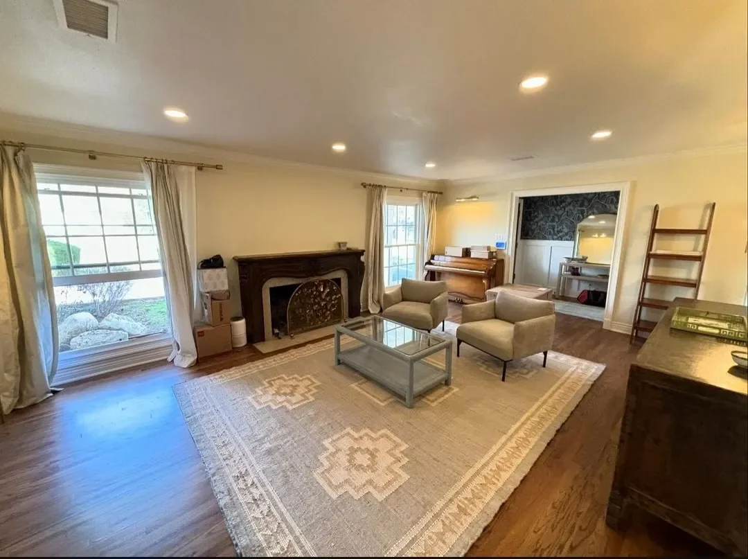 Living room with large window, two beige armchairs, glass coffee table, fireplace, upright piano, arranged on a beige patterned rug, hardwood flooring, and soft overhead lighting.