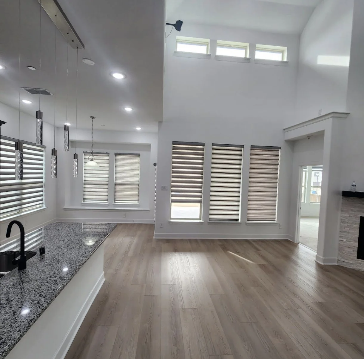 Empty modern living space with hardwood floors, large windows with shades, white walls, recessed and hanging lighting, and a granite kitchen counter with a black sink.