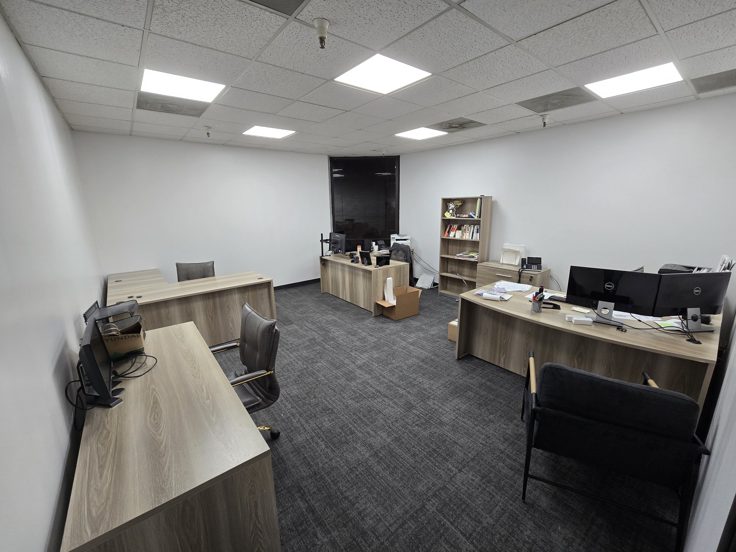 Empty office space with four desks, two with computer monitors, and a black armchair. There are some scattered papers and office supplies, a bookshelf with books, and a window with closed blinds. The ceiling has fluorescent lights and suspended tiles