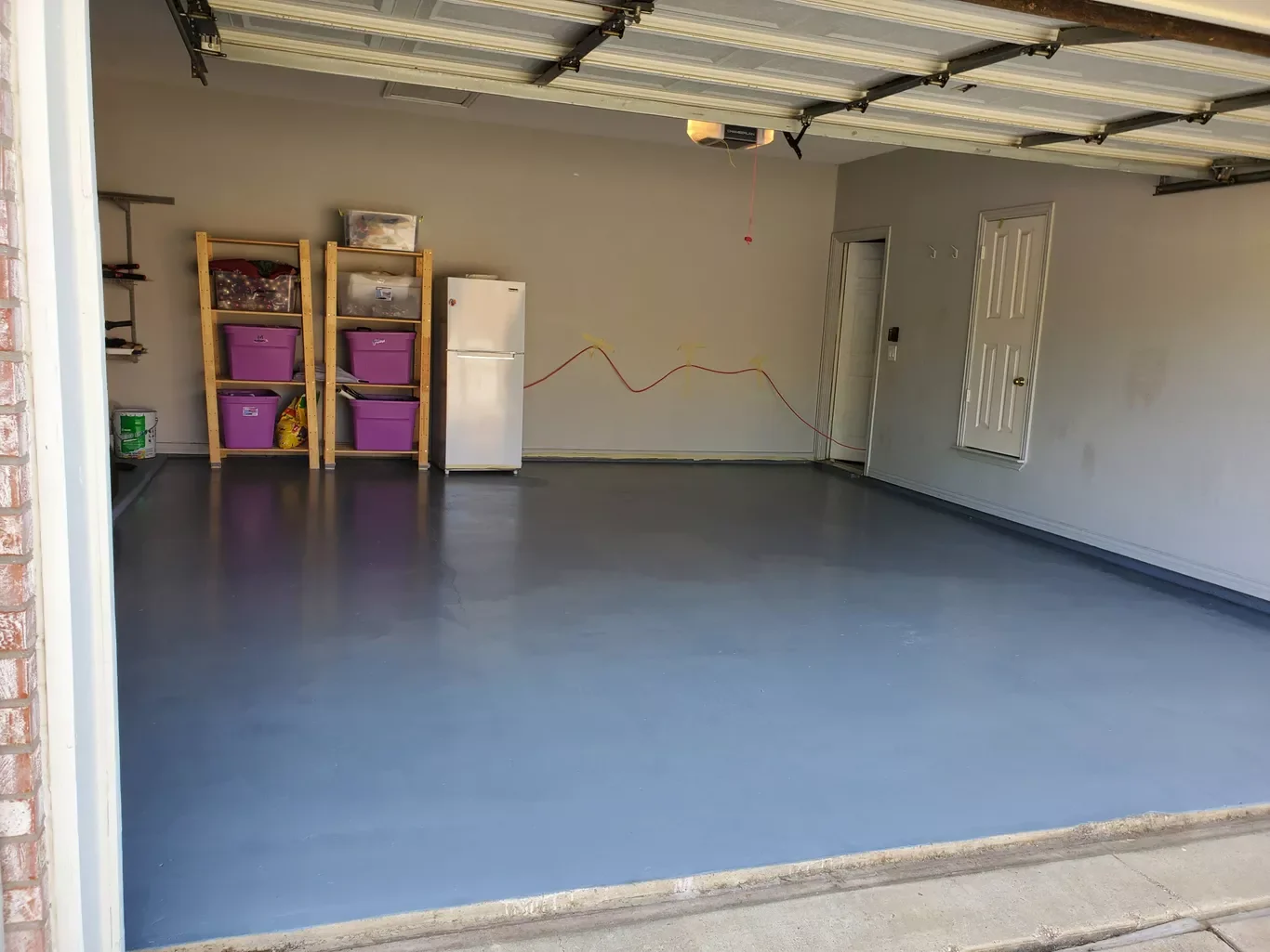 Empty garage with freshly painted blue floor, storage shelves with purple bins, small refrigerator, and garage door open.