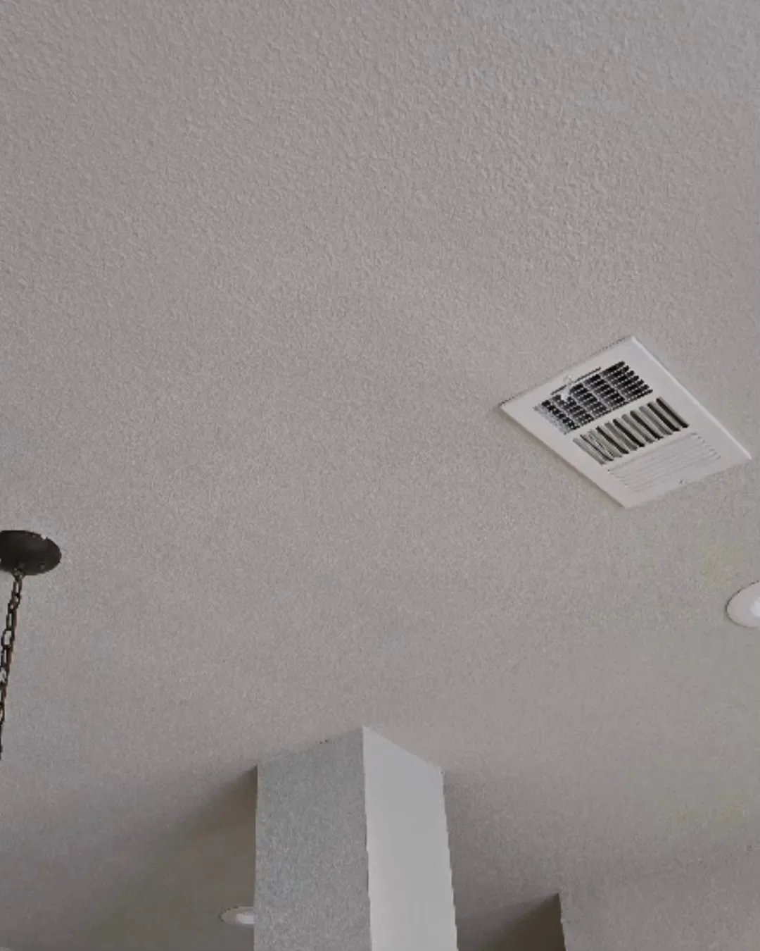 Ceiling with a vent and a light fixture, partially visible wall, and a hanging chain light fixture.