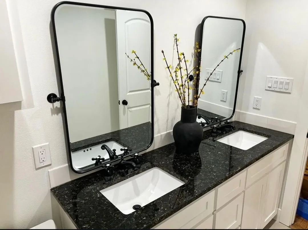 Double bathroom vanity with black granite countertop, two rectangular sinks, black fixtures, two rectangular mirrors with black frames, a black vase with yellow flowering branches, and wall-mounted switches and outlets.