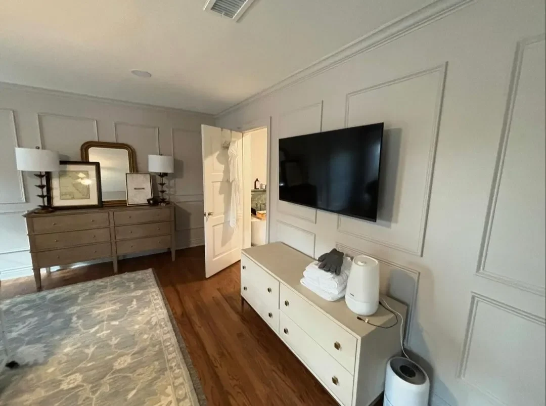Living room with white wall paneling, flat-screen TV mounted on wall, white dresser with towels and air purifier, hardwood floor, decorative mirror, and framed artwork on dresser
