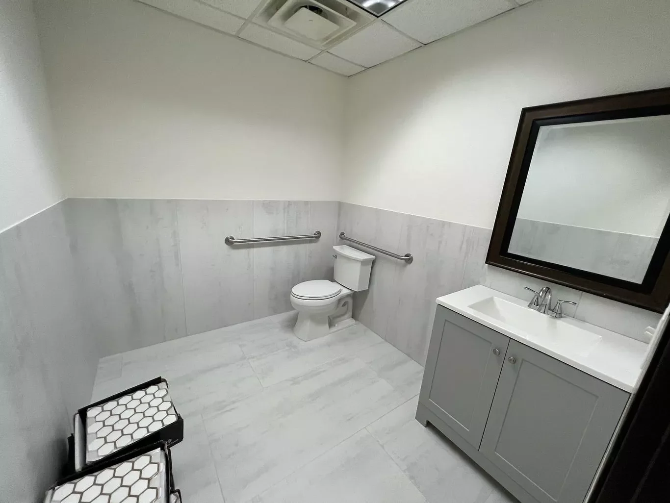 A clean and empty commercial bathroom with a white toilet, a gray vanity with a sink, a large mirror, and handrails on the wall, with a small black and white hexagonal patterned stool in the corner.