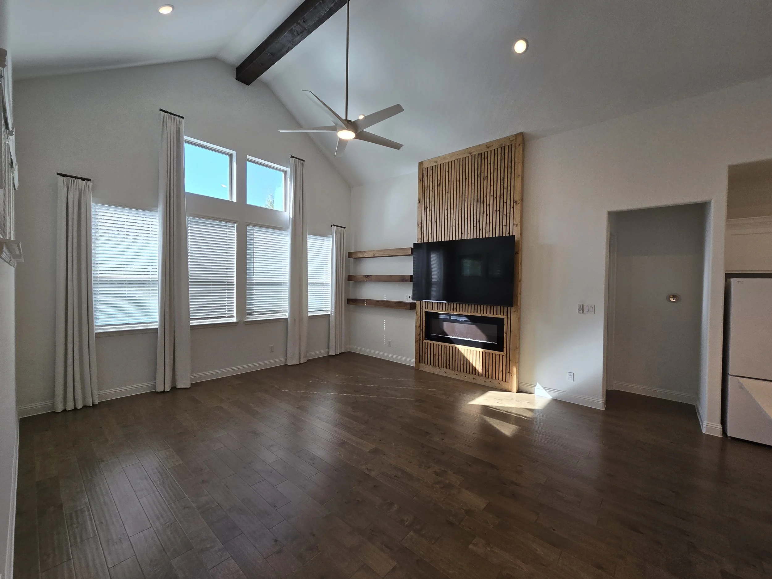 Empty living room with large windows, white walls, hardwood flooring, and a ceiling fan. There is a mounted black TV on a wooden accent wall with a built-in fireplace below, and two wooden shelves to the right of the TV.