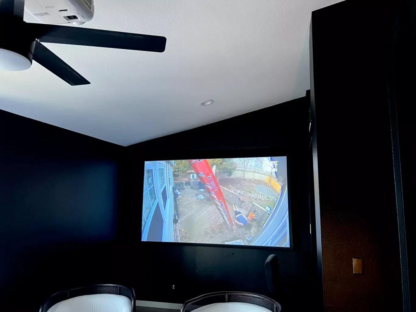 Interior of a dark room with a ceiling fan and a large screen showing a backyard construction site with a red ladder and outdoor furniture.