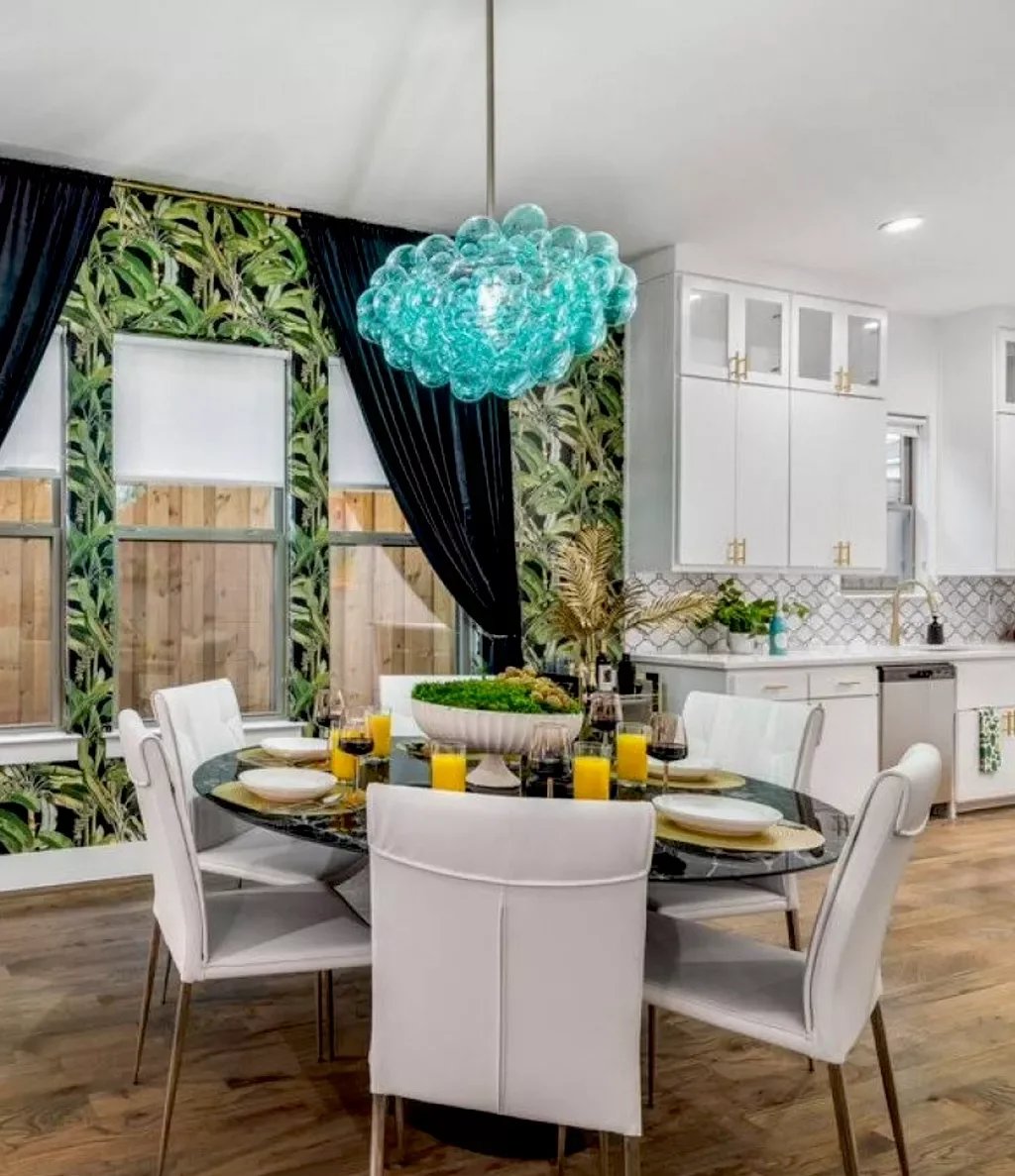 Dining room with a round table set with white plates, yellow drinks, wine glasses, and a green centerpiece. There are six white chairs around the table. The room has a tropical wallpaper on one wall, large windows with black curtains, and a modern ch
