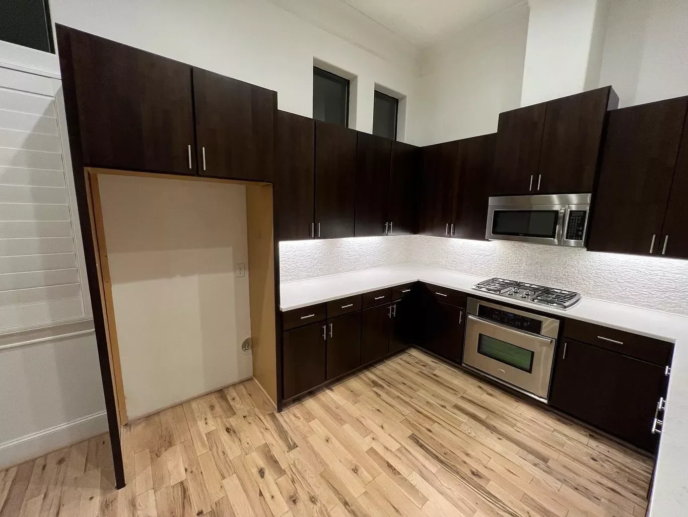 Modern kitchen with dark upper and lower cabinets, stainless steel microwave, oven, and gas stove, white textured backsplash, and wood flooring.