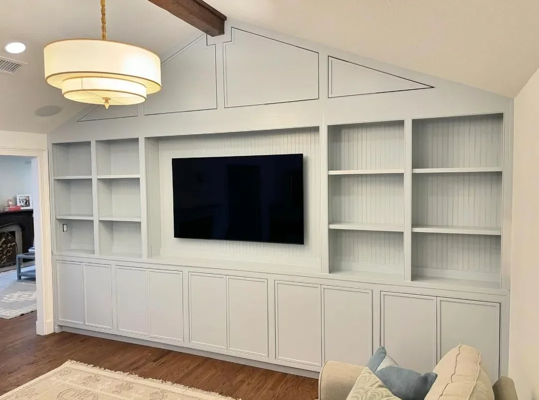 Living room with a TV mounted on a built-in white wall unit with shelves and cabinets, a beige sofa with pillows, hardwood floor, and a ceiling light fixture.