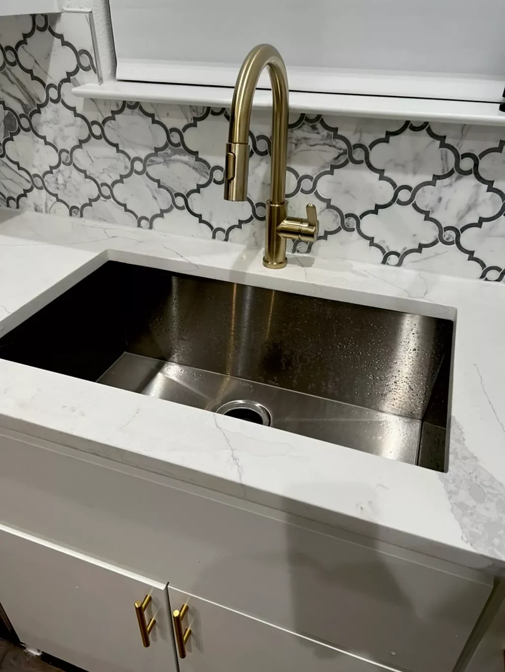 Kitchen sink with a gold faucet, white marble countertop, and patterned tile backsplash.