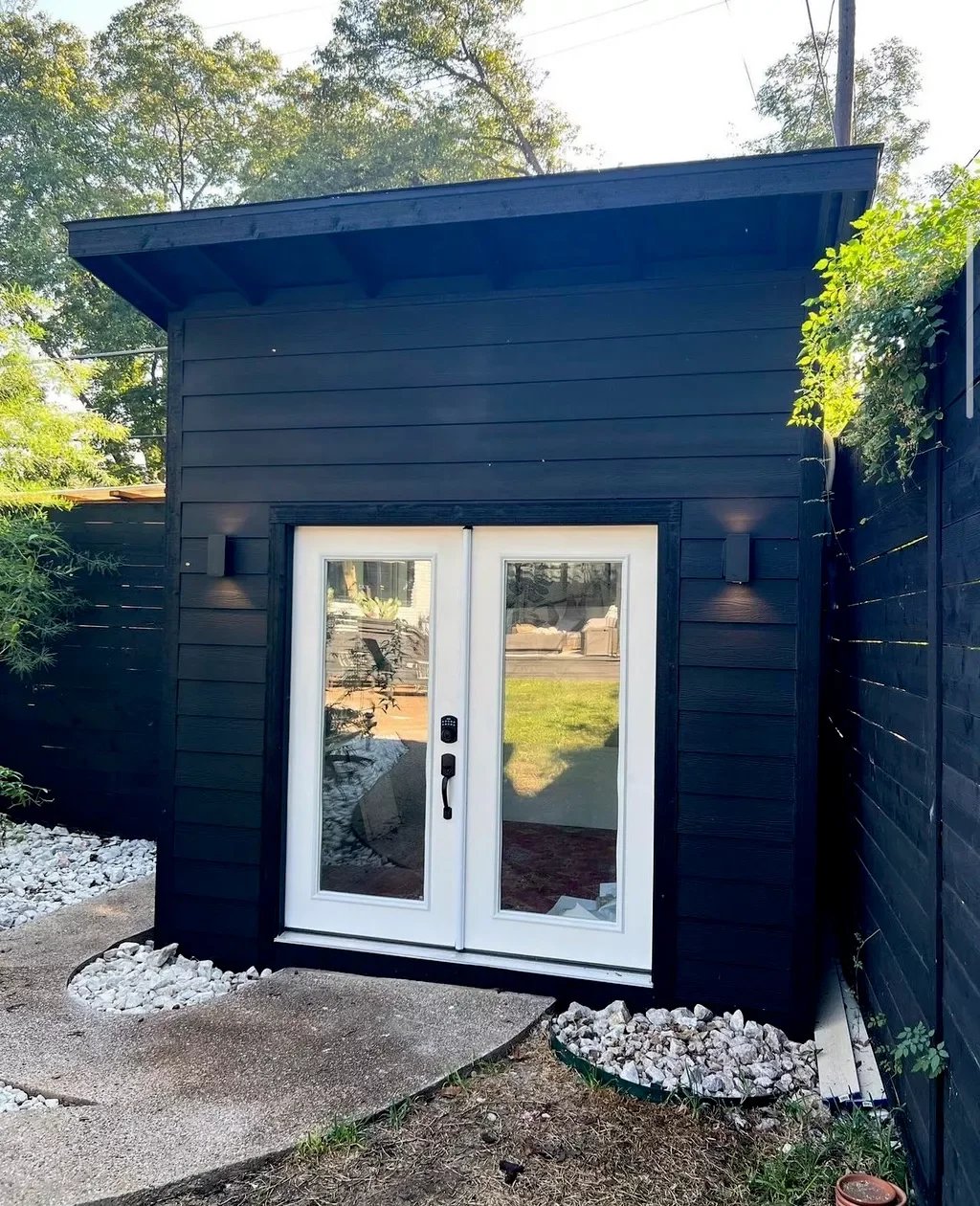 A small black shed with a sloped roof and double glass doors, surrounded by a landscaped yard with white rocks and a concrete pathway.