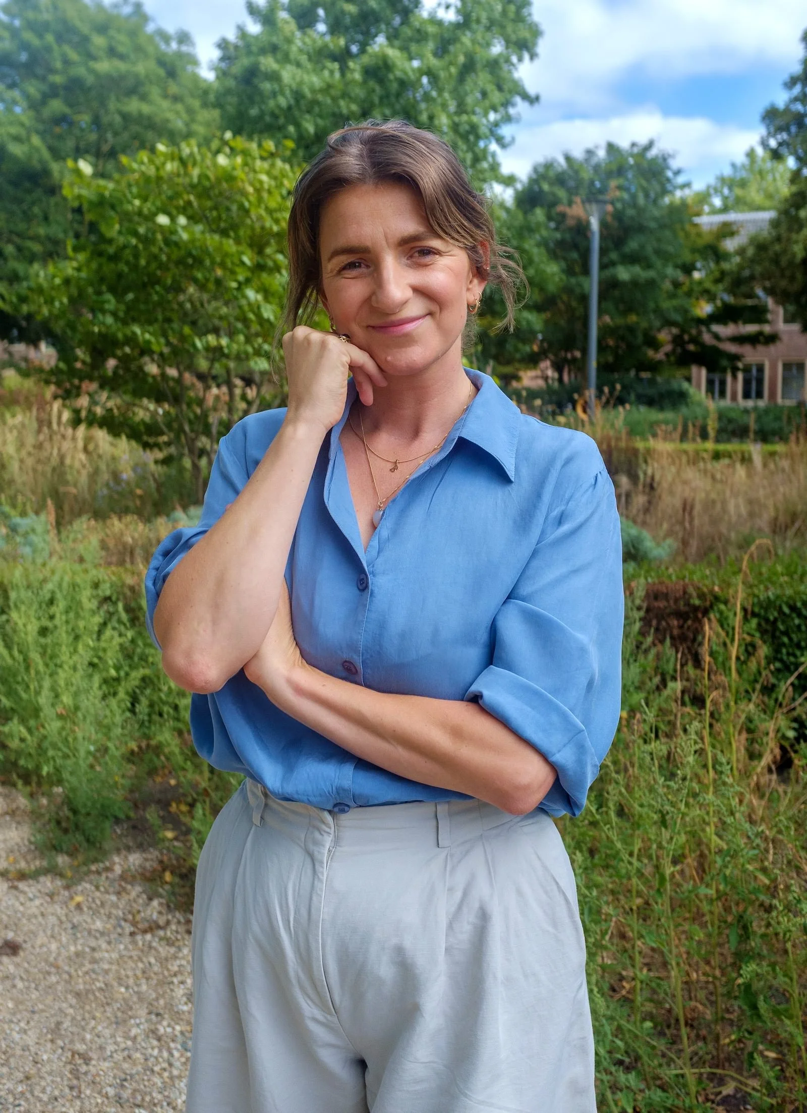 Een vrouw met korte bruin haar, die een glimlach vertoont, draagt een blauwe blouse en staat buiten in een groene omgeving met bomen en een partly cloudy sky.