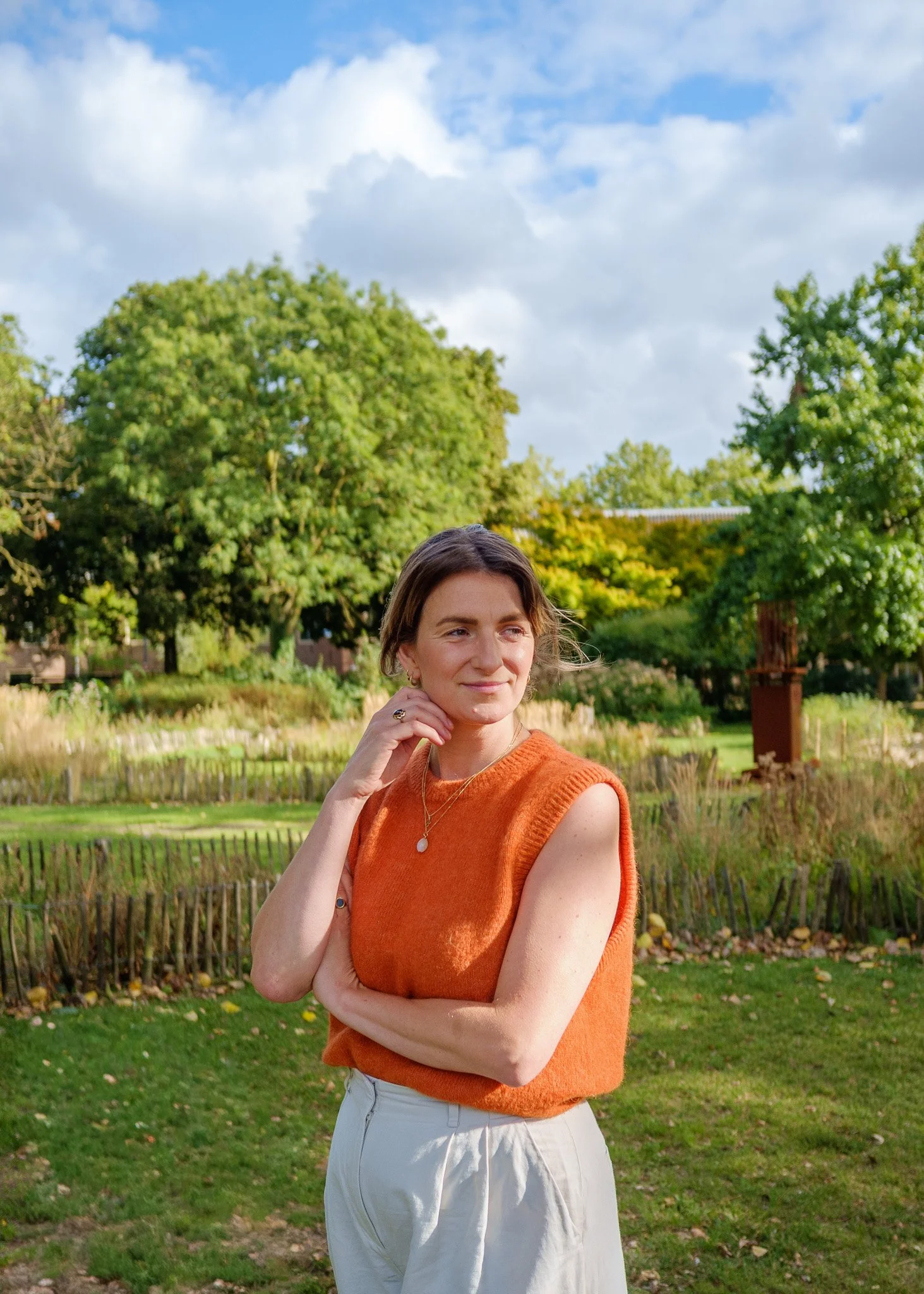een jonge vrouw met bruin haar draagt een oranje top en witte broek, staat buiten in een groene park met bomen en een houten bank op de achtergrond, onder een bewolkte blauwe lucht.